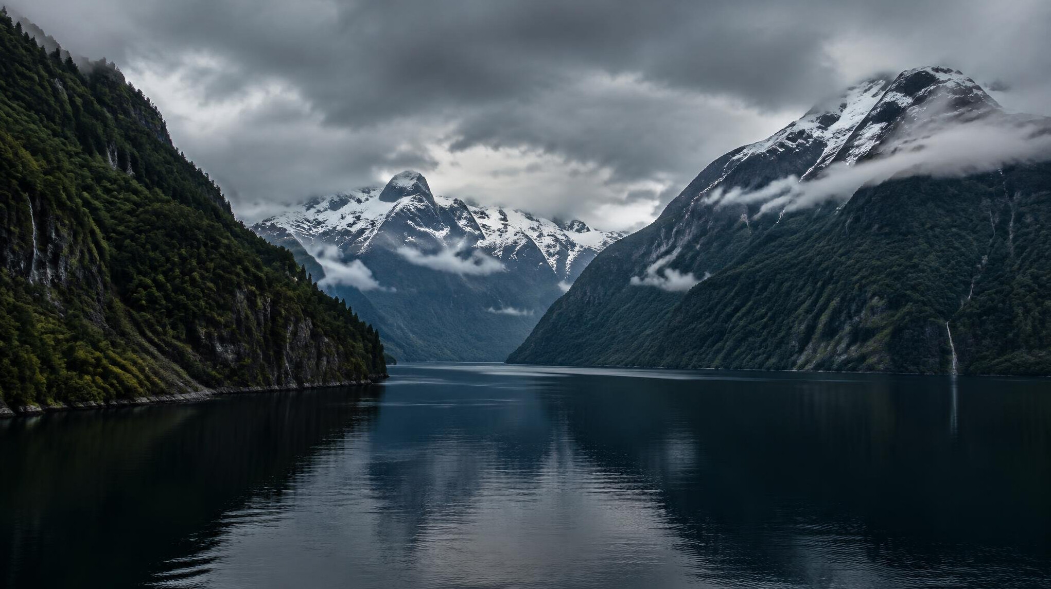 Patagonian fjord with dark water and snowy mountains under clouds
